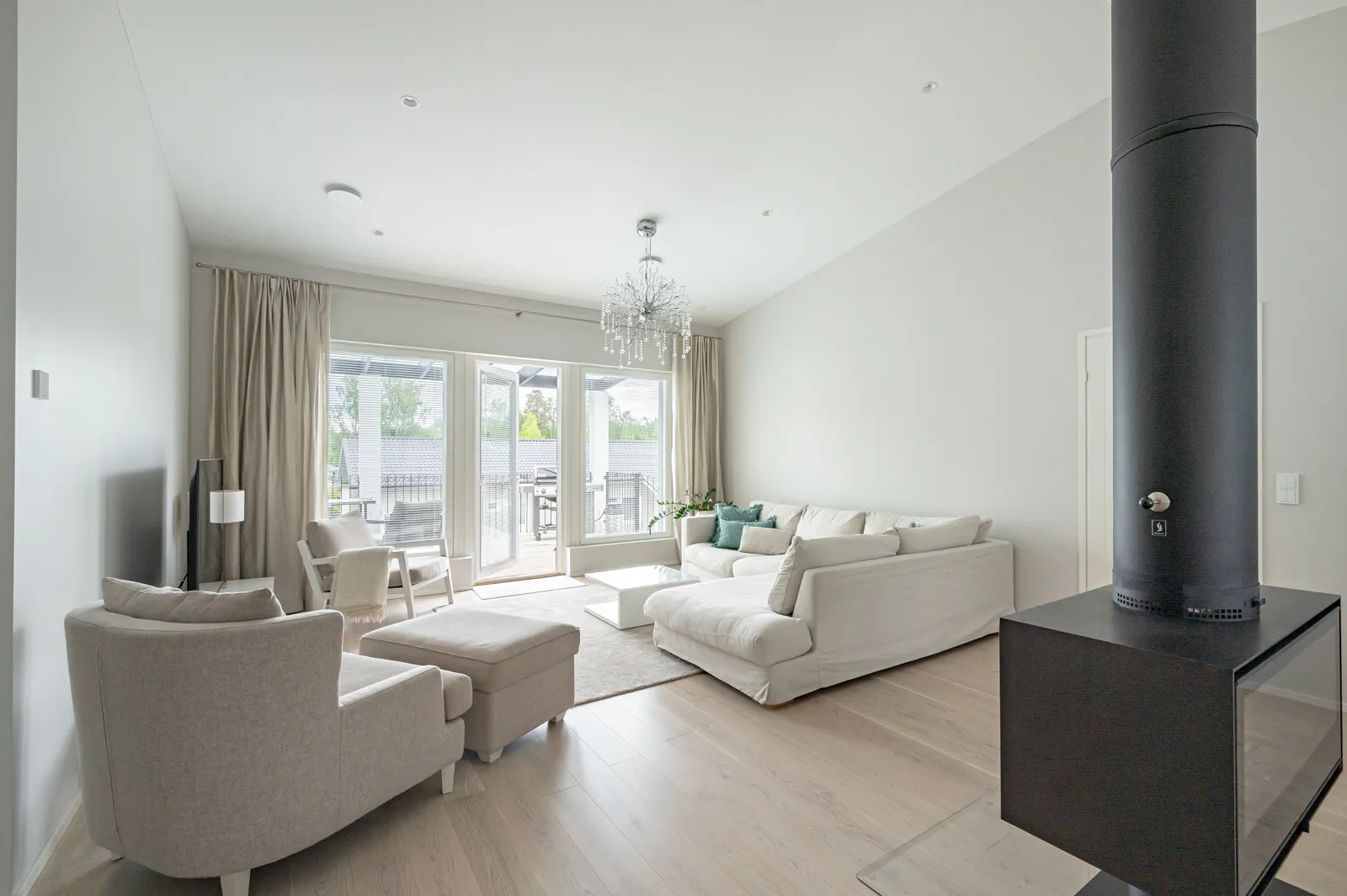 Vaaleassa olohuoneessa on suuri valkoinen sohva, harmaa nojatuoli jalkarahilla, moderni takka ja kristallikruunu. Suuret ikkunat tuovat runsaasti luonnonvaloa.