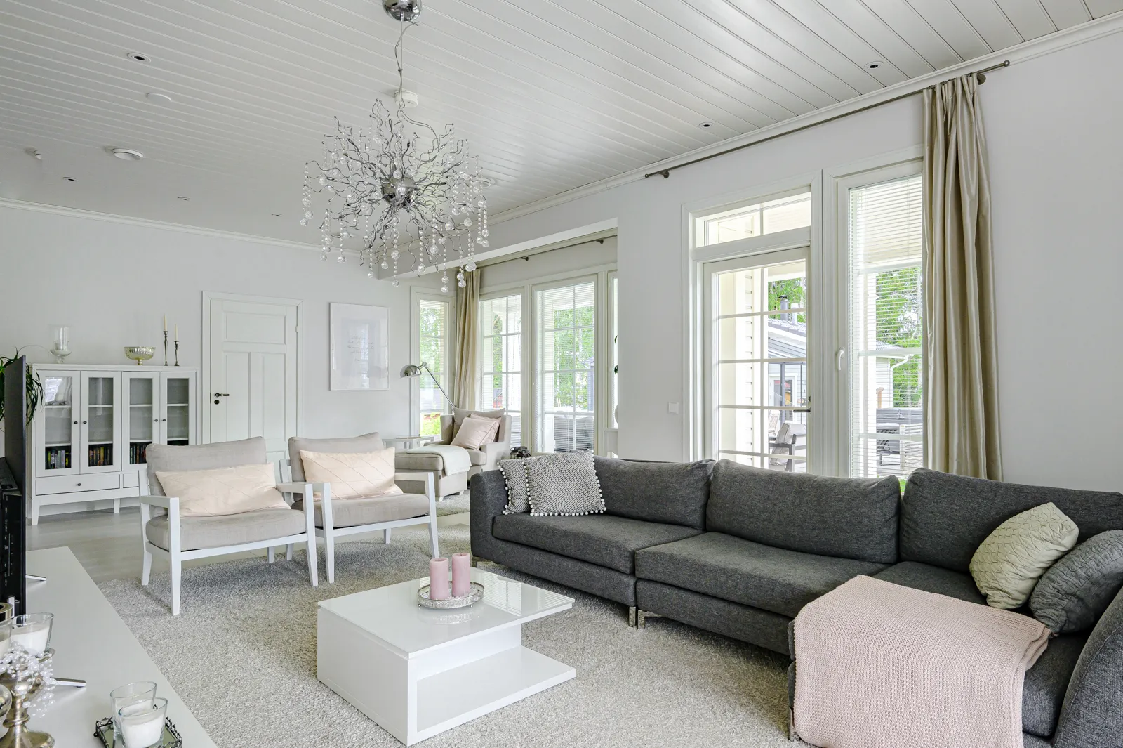 Vaaleassa olohuoneessa on harmaa sohva, valkoinen sohvapöytä, kaksi harmaata nojatuolia ja kristallikruunu.