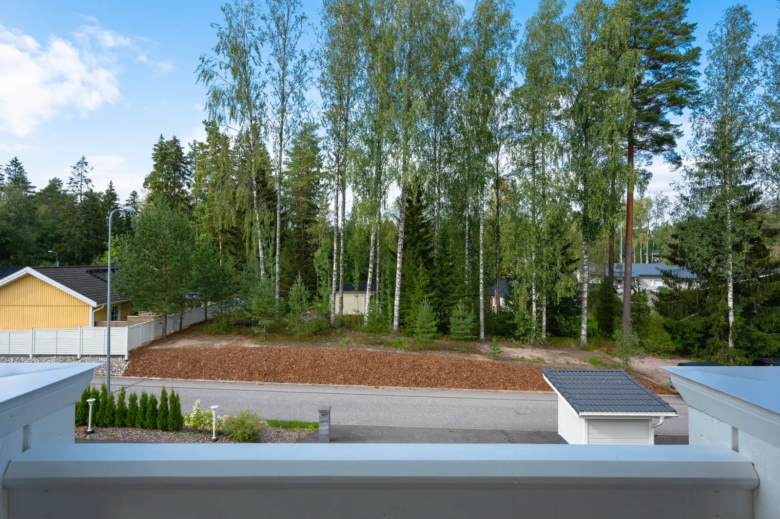Näkymä parvekkeelta: puita, taloja ja asfalttitie. Aurinkoinen sää.