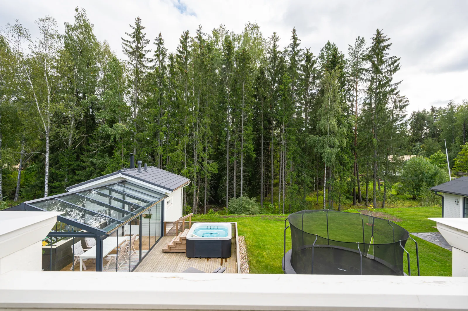 Näkymä parvekkeelta, jossa näkyy lasitettu terassi, poreallas, trampoliini ja vehreä metsäinen maisema.