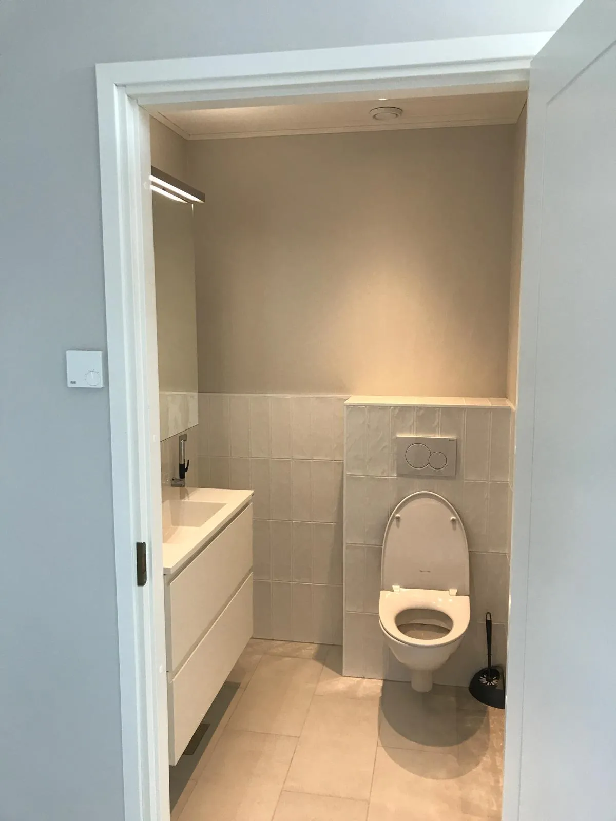 Pieni kylpyhuone, jossa on valkoinen pesuallas, seinään kiinnitetty WC ja beigejä laattoja. Huone on valaistu ylhäältä.