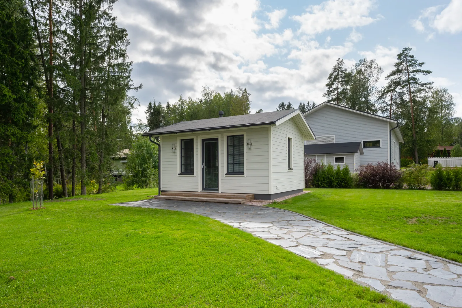 Pieni, valkoinen piharakennus kivipolun päässä, ympärillä vihreä nurmikko ja puita, taustalla suurempi harmaa talo pilvisen taivaan alla.