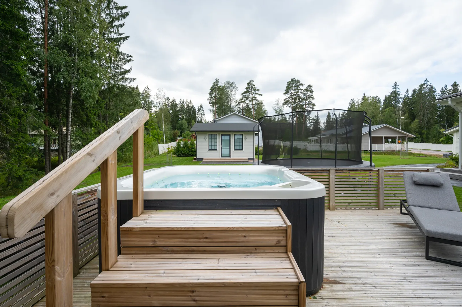 Ulkona on poreallas, puinen terassi, portaat, lepotuoli ja taustalla näkyy trampoliini ja rakennuksia pilvisen sään alla.