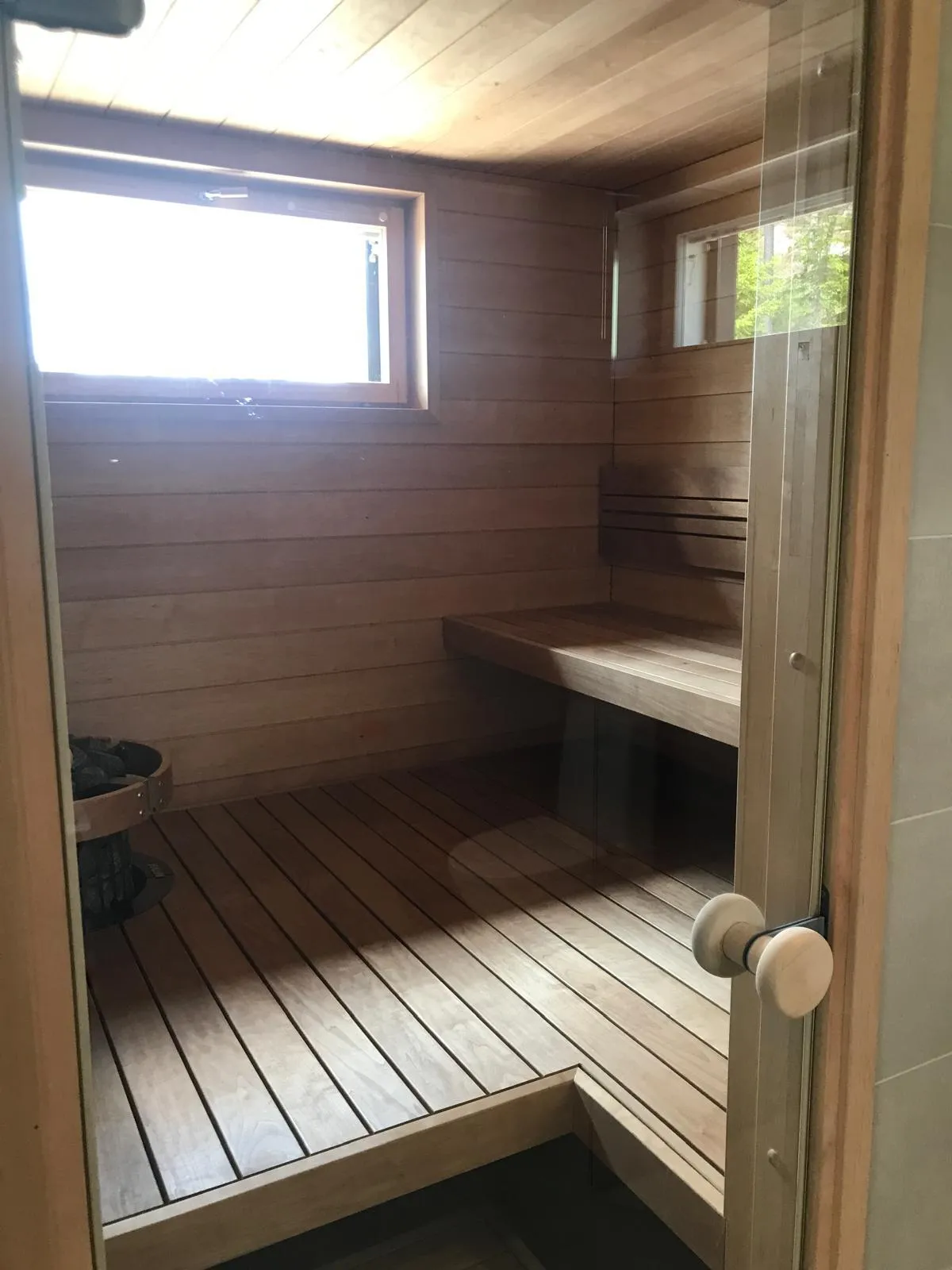 Puinen sauna, jossa on penkki, ikkuna ja lasiovi, valaistus on kirkas.