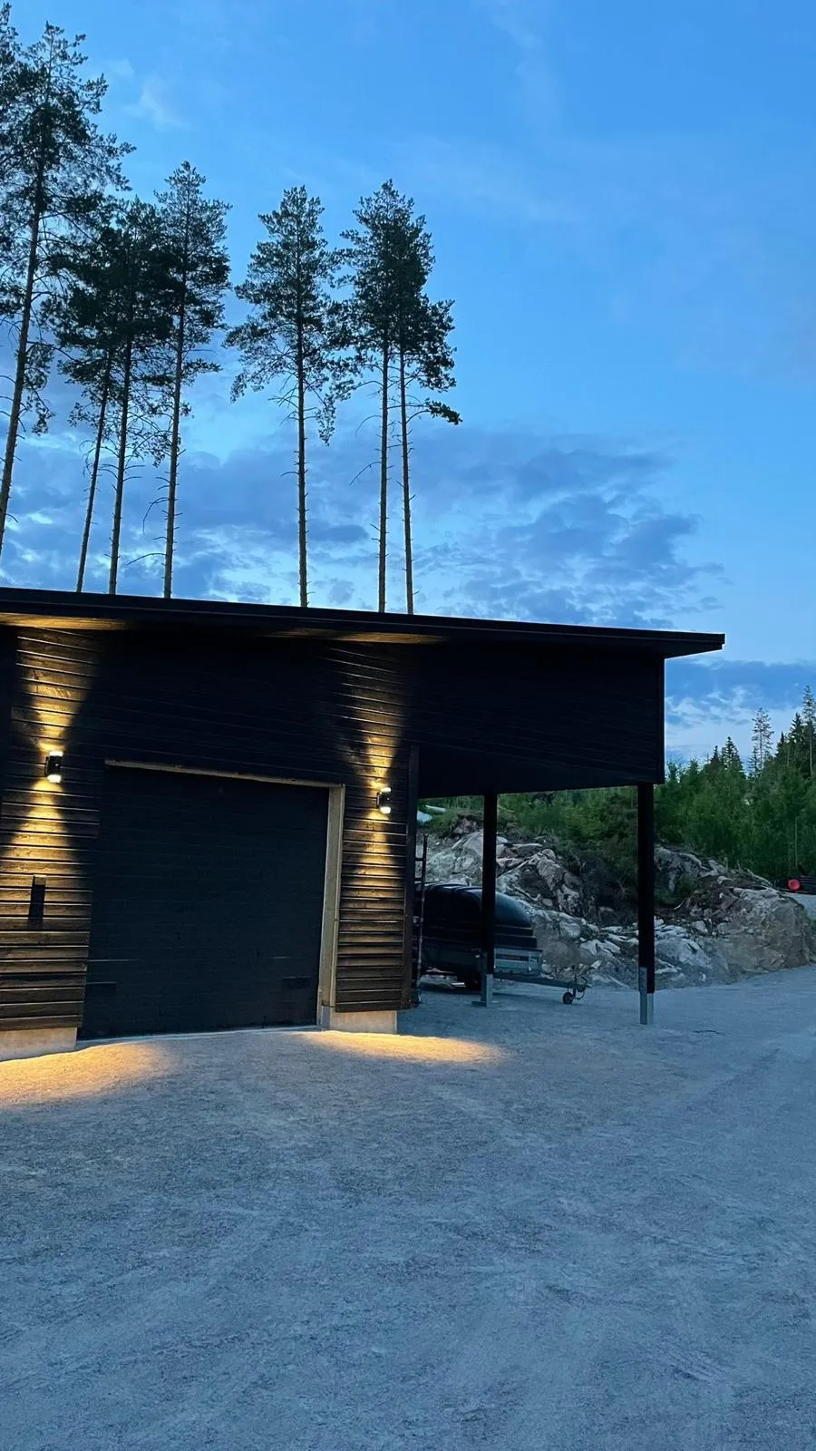 Tumma puurakennus, jossa on autotalli ja katos, valaistu ulkovaloilla, taustalla puita ja sininen taivas.
