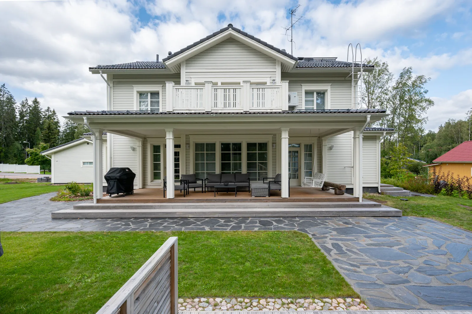Kaksikerroksinen, vaaleanharmaa puutalo, jossa on suuri katettu terassi, kivipolku ja vihreä nurmikko aurinkoisena päivänä.