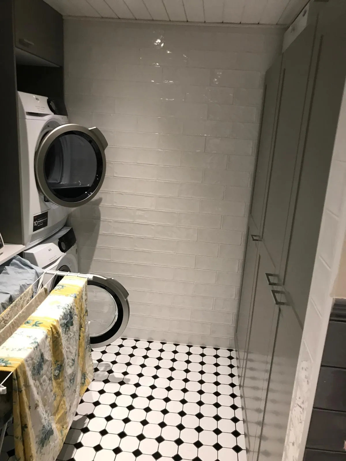 Pesukoneet, kuivausteline ja mustavalkoinen lattia pesuhuoneessa, jossa on valkoiset seinälaatat ja harmaa kaappi.