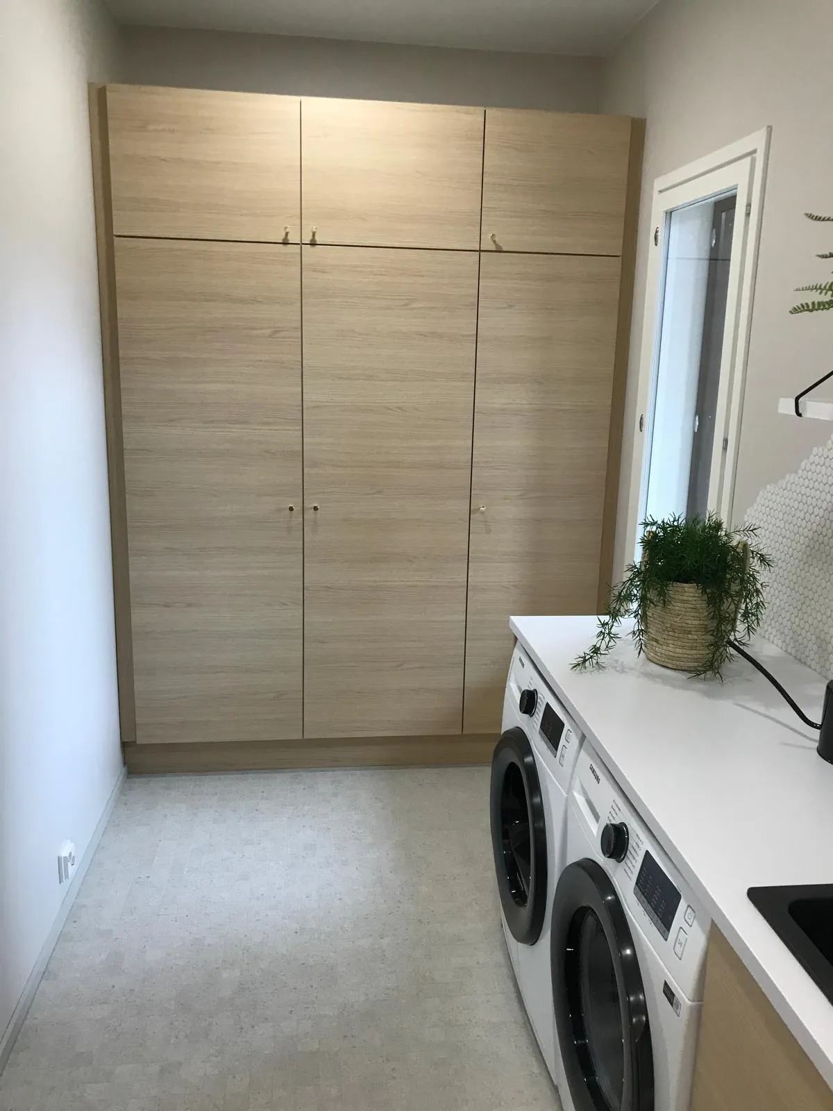 Vaaleassa kodinhoitohuoneessa on kolme suurta puukaappia, valkoinen pesukone ja kuivausrumpu sekä viherkasvi valkoisella tasolla.