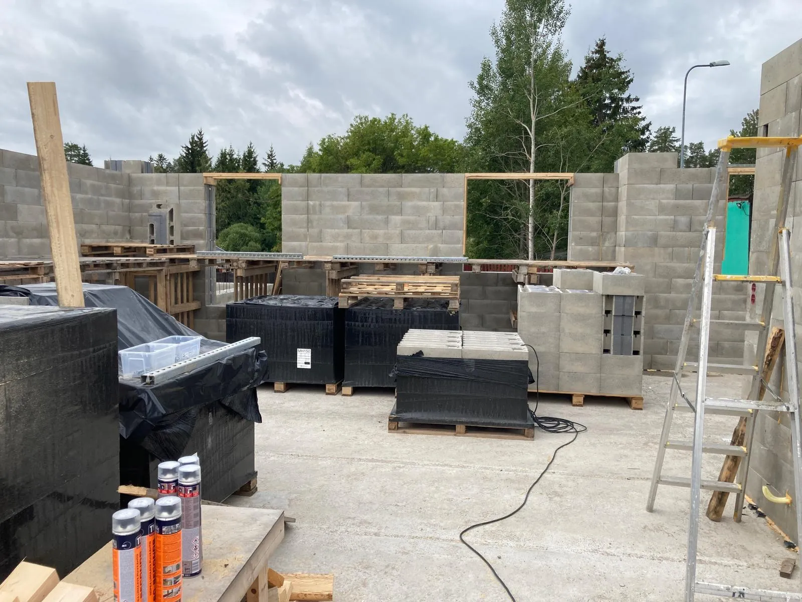 Rakennustyömaa, jossa on harmaita betoniharkkoja, puupaletteja, mustia paketteja ja alumiinitikkaat pilvisessä säässä.
