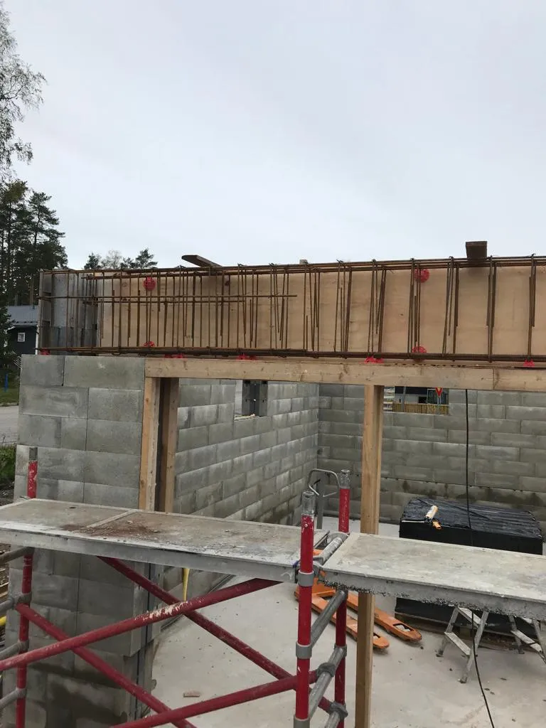 Rakennustyömaa, jossa on harmaita betoniharkkoja, puinen kehys ja punainen rakennusteline, valoisassa säässä.