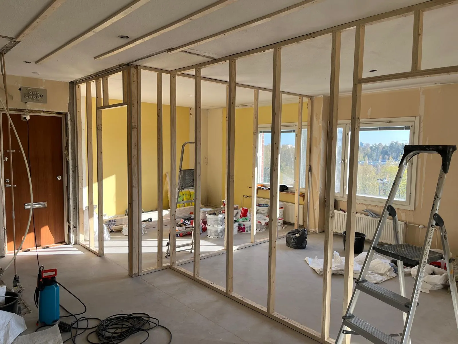 Rakennustyömaa, jossa puurunkoisia seiniä, maalipurkkeja, tikkaita ja siivousvälineitä valoisassa huoneessa.