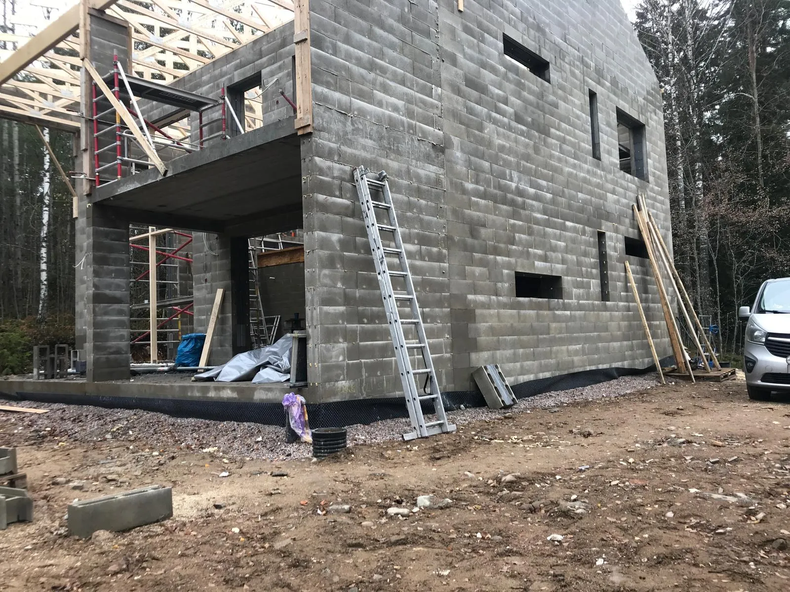 Kaksikerroksinen harmaa harkkorakennus rakenteilla, puinen kattorakenne näkyvissä, alumiinitikkaat nojaavat seinään, maassa rakennustarvikkeita ja auto osittain näkyvissä.