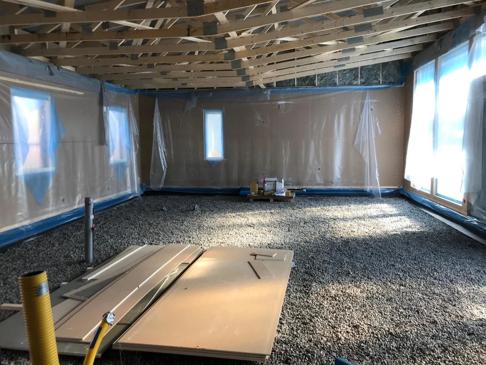 Rakenteilla oleva huone, jossa on kivipohja, muovilla peitetyt seinät ja puinen kattorakenne. Valoisa ja avara tila.