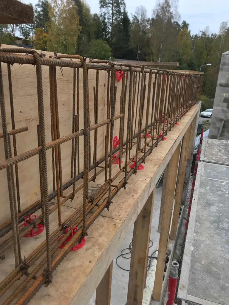 Rakennustyömaa, jossa näkyy raudoitus ja puumuotti, punaiset muoviset välikappaleet ja taustalla puita.