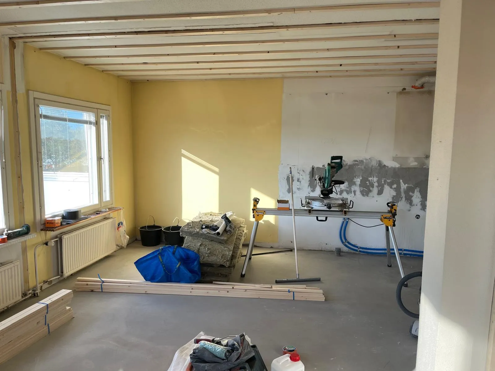 Huone remontissa, jossa on ikkuna, puutavaraa, eristystä, sahauspöytä ja työkaluja. Seinät ovat osittain maalattuja, katossa on puurimoitus.
