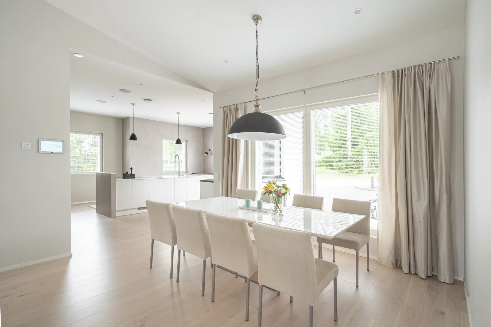 Vaaleassa ruokailutilassa on valkoinen pöytä, kuusi beigeä tuolia, suuri ikkuna verhoineen ja musta riippuvalaisin.