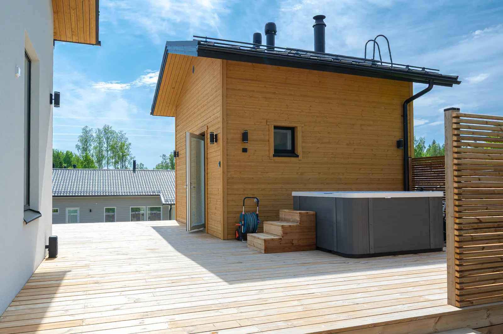 Puinen sauna, poreallas ja terassi aurinkoisena päivänä, sininen taivas.