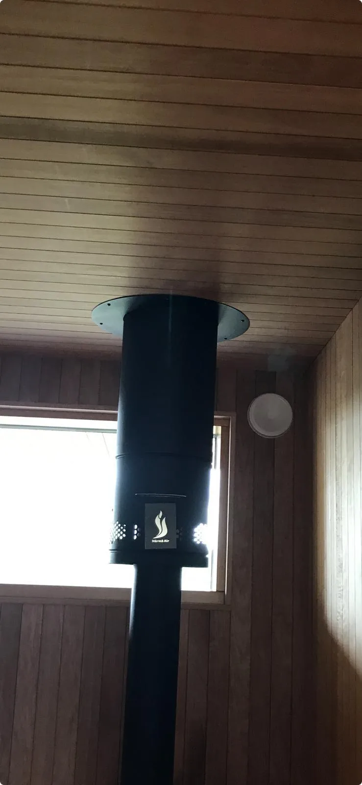 Musta savupiippu puupaneloidussa saunassa, jossa on ikkuna ja valaisin.