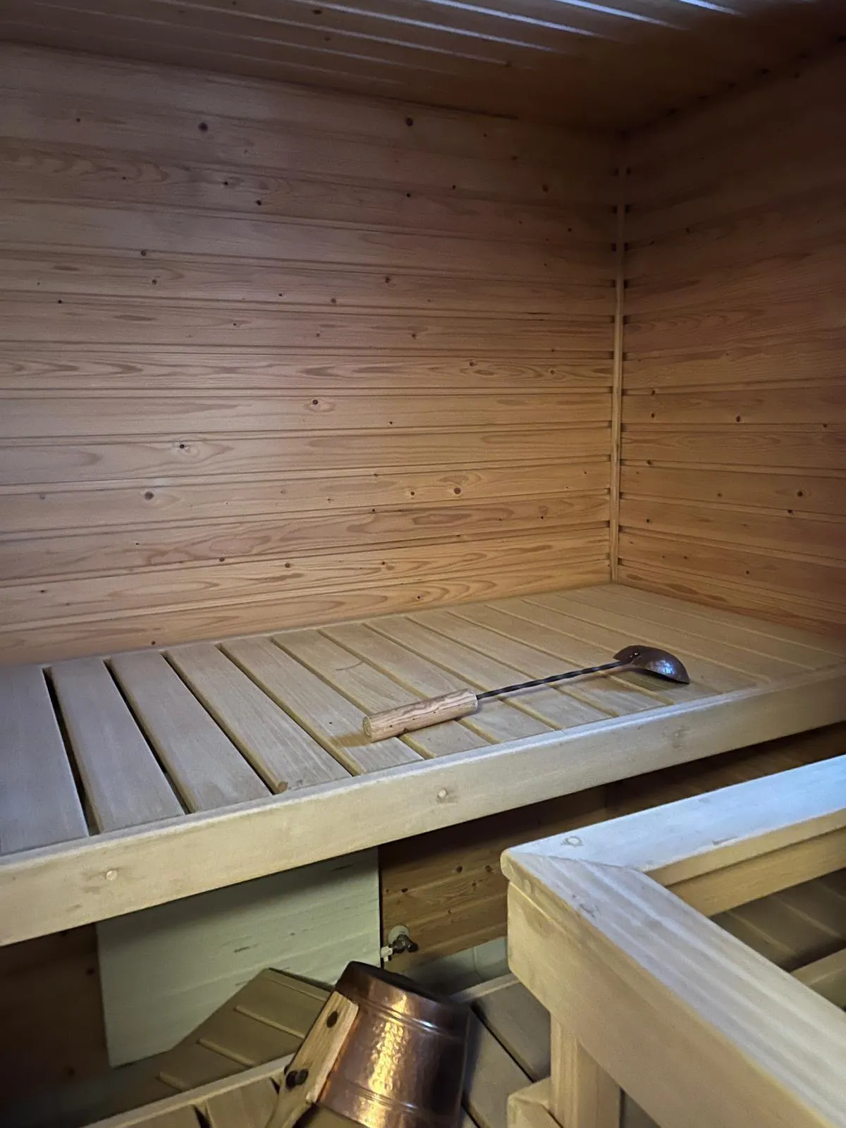 Sauna puupaneeleilla, kauha penkillä ja kupariämpäri lattialla, lämmin valaistus.