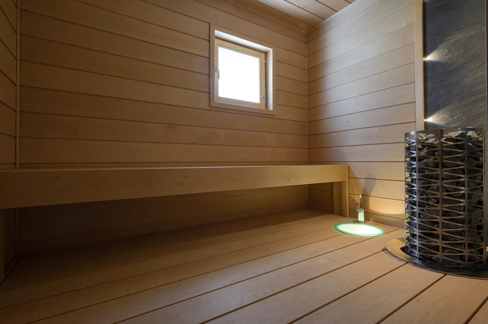 Puinen sauna, jossa on ikkuna, penkki ja kivikiuas, valaistus luo lämpimän tunnelman.