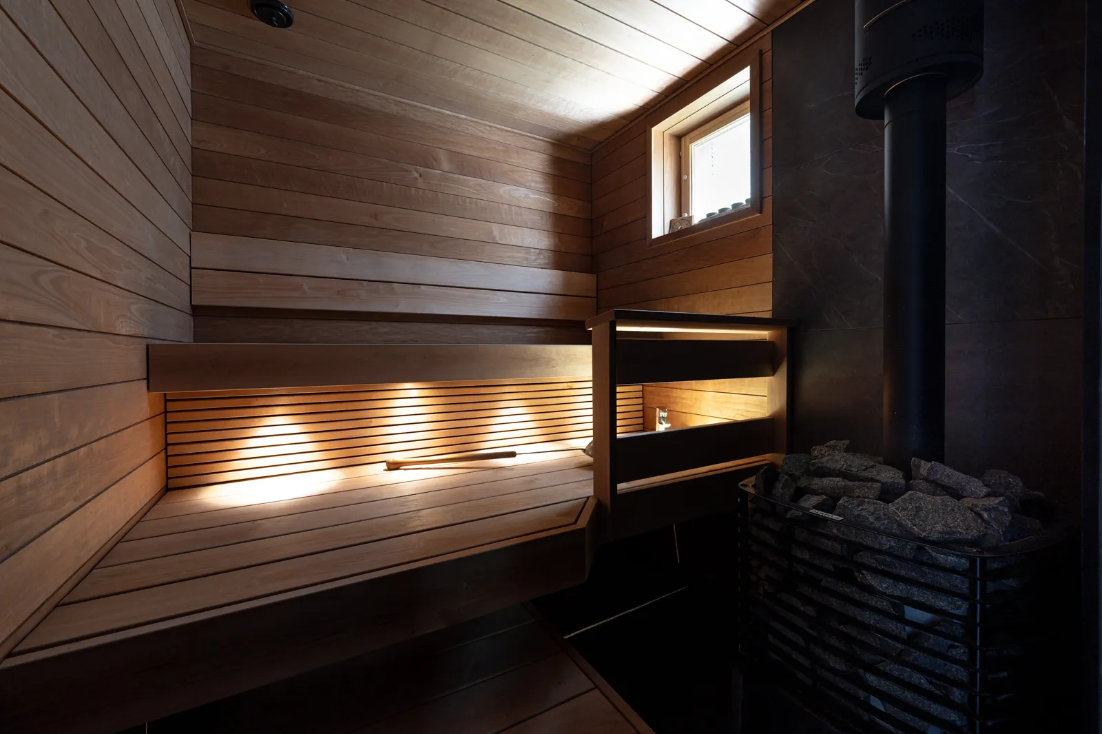 Sauna, jossa on puupanelointi, penkit, ikkuna ja kiuas kivillä, luoden lämpimän ja rentouttavan tunnelman.