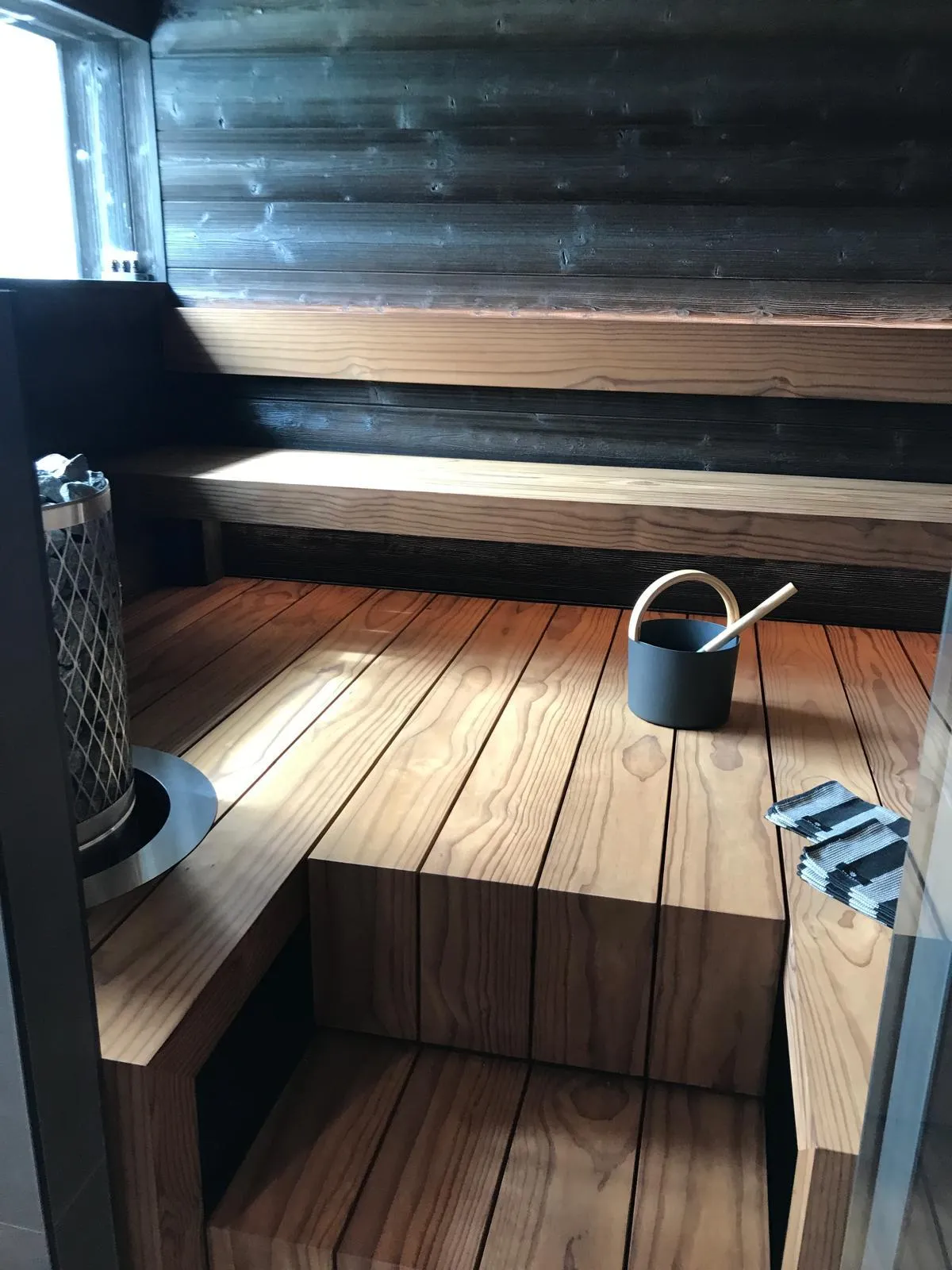 Sauna, jossa on puupaneelit, penkit, kiuas, kauha ja laudeliinat. Valoisa ja lämmin tunnelma.