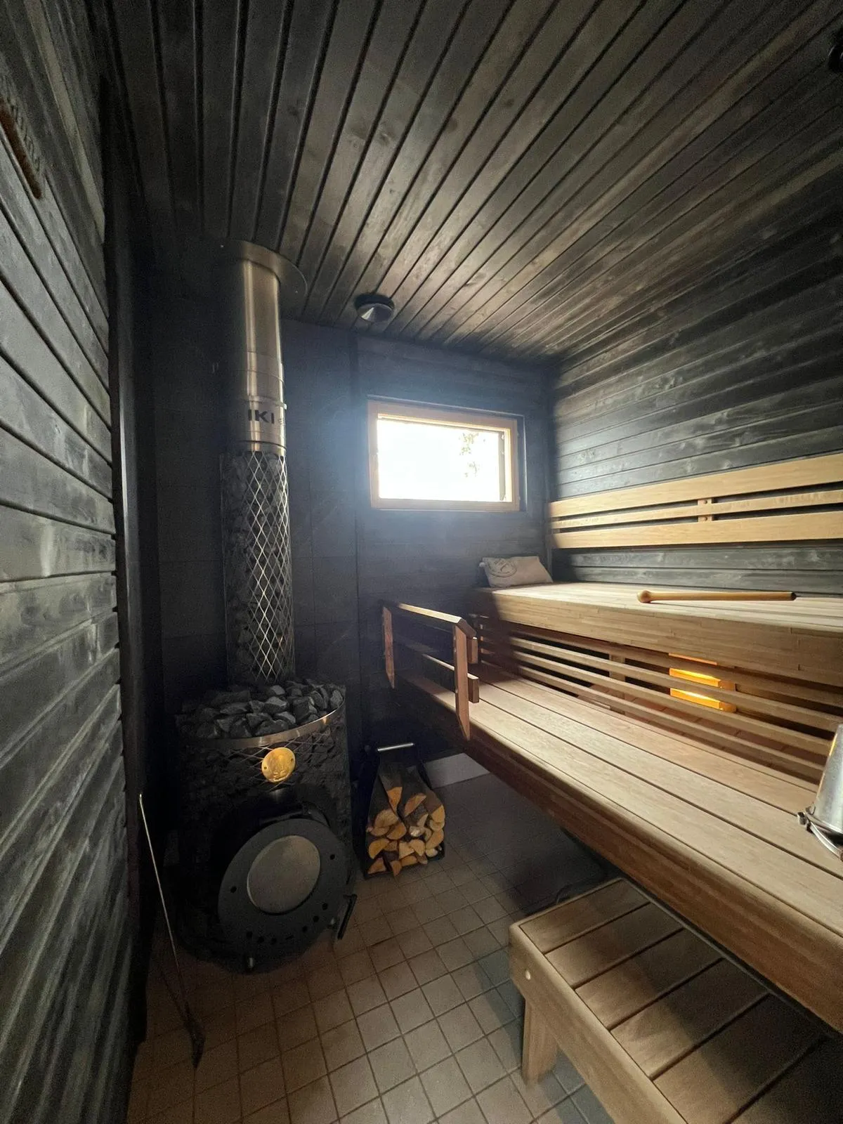 Sauna tummalla puupaneloinnilla, puukiuas kivillä ja puulauteet. Ikkuna tuo valoa tilaan.