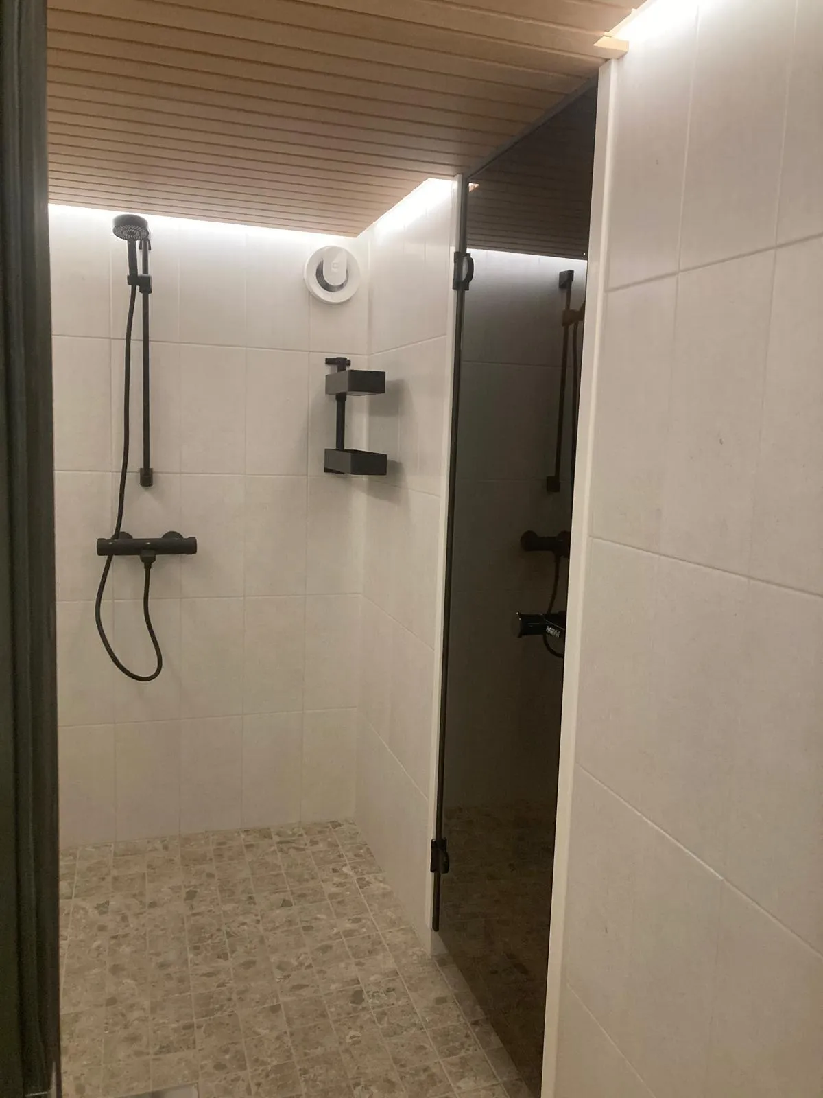 Kylpyhuoneen sisustus, jossa on suihku, laattalattia ja puinen katto. Suihkussa on musta suihkupää ja hylly. Seinät on laatoitettu valkoisella laatalla.