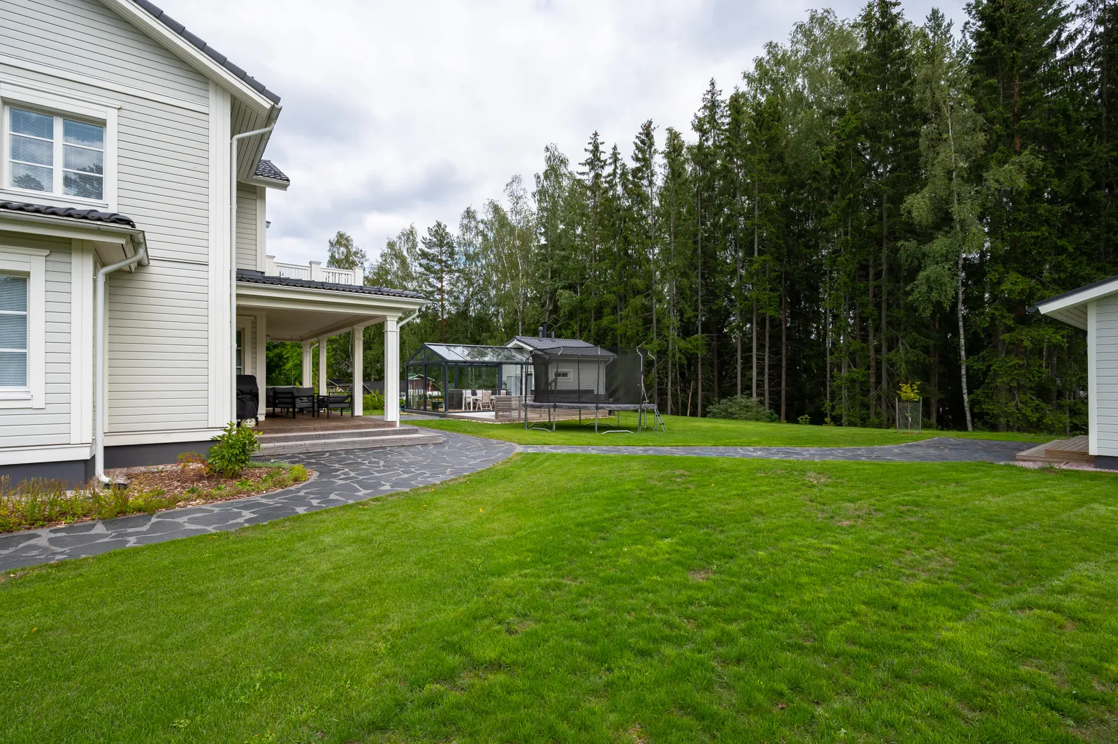 Kuvassa on valoisa ja vehreä takapiha, jossa on nurmikko, polku, talo ja metsää taustalla.