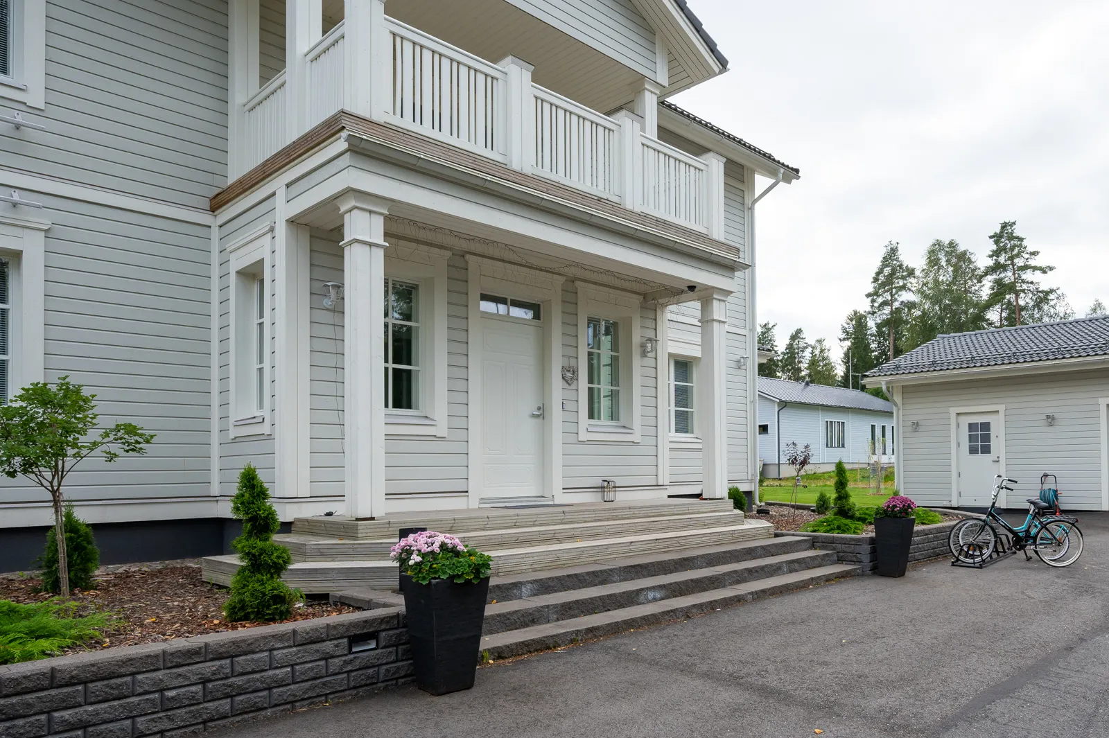 Vaaleanharmaa kaksikerroksinen talo, jossa on valkoinen kuisti, kiviset portaat ja mustat kukkaruukut, sekä polkupyörä autotallin edessä.