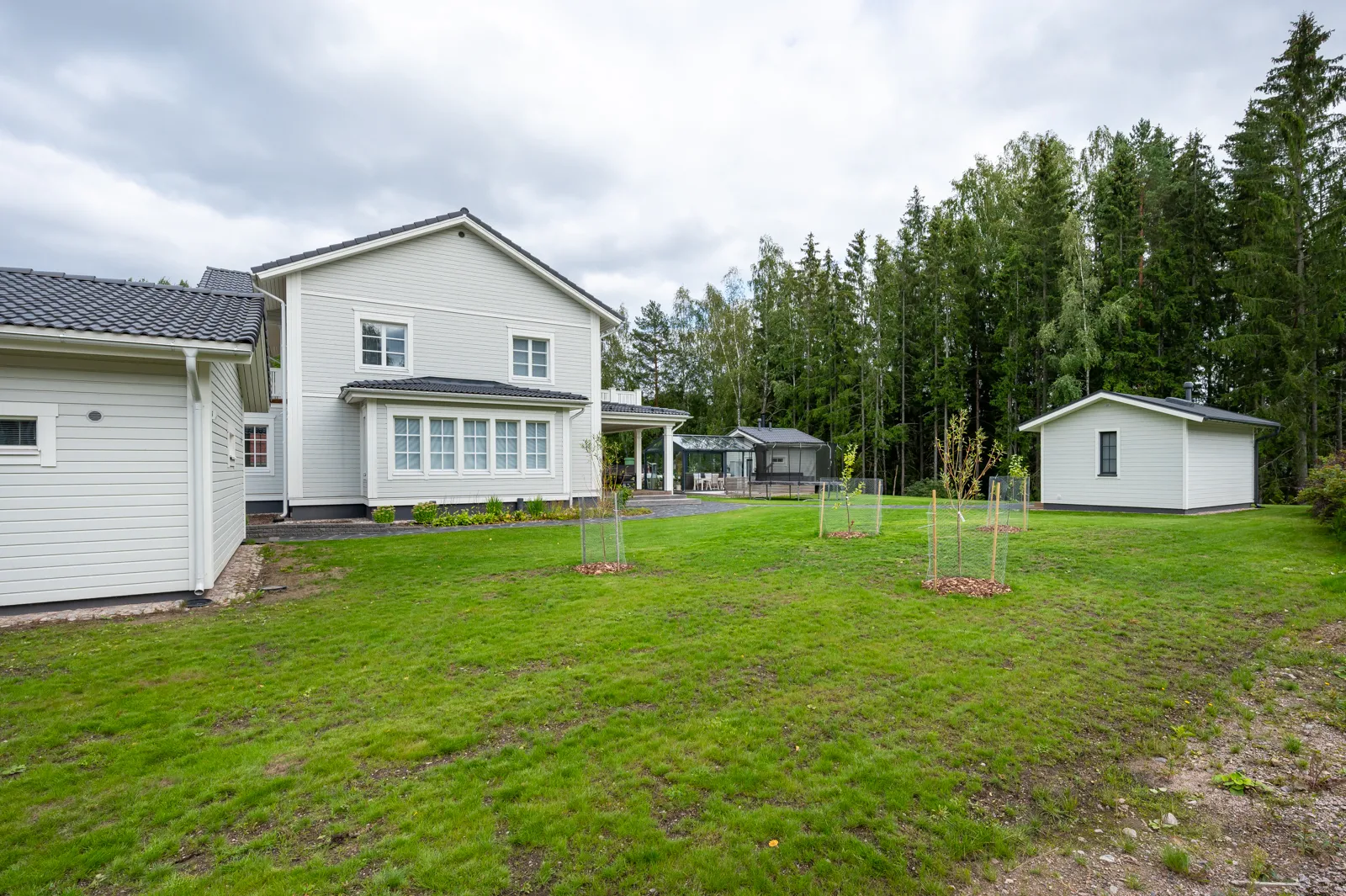 Vaaleanharmaa kaksikerroksinen talo, jossa on tumma tiilikatto, vihreä nurmikko ja puita pilvisen taivaan alla.