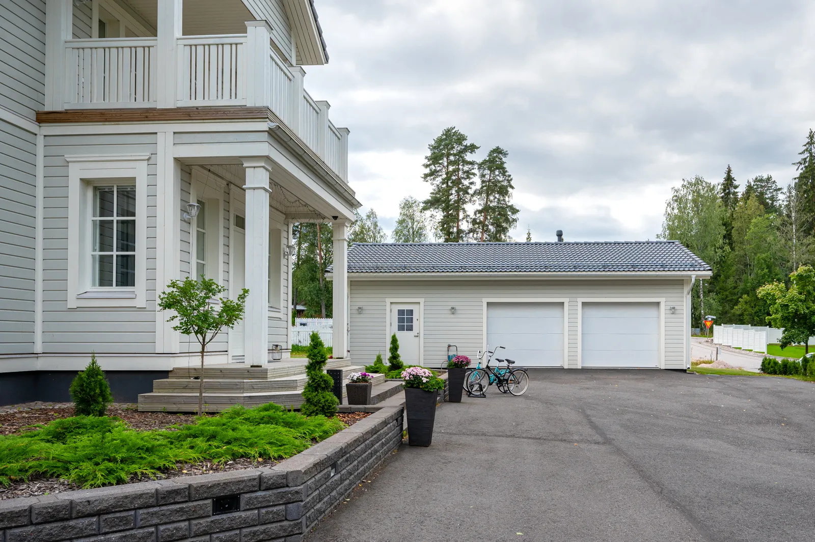 Vaaleanharmaa talo, jossa on valkoiset koristeet, parveke, autotalli, kaksi polkupyörää ja vihreä piha pilvisen taivaan alla.