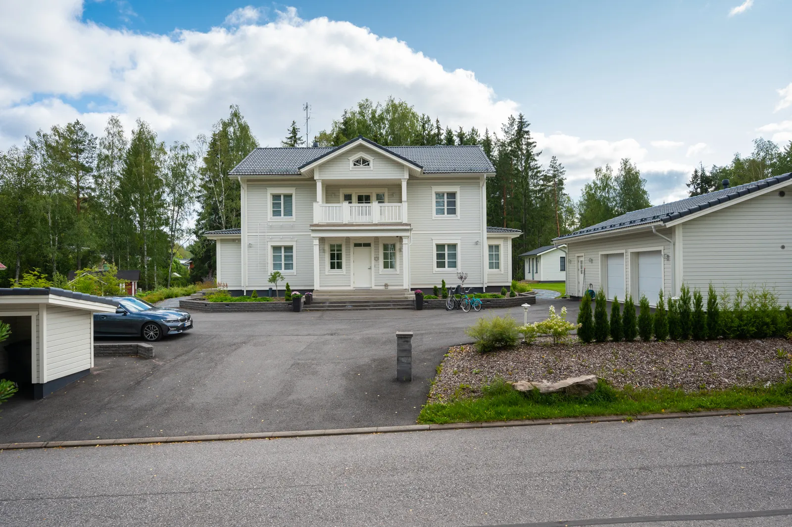 Vaaleanharmaa kaksikerroksinen talo, jossa on valkoinen parveke ja tumma katto, sijaitsee asfaltoidulla pihalla, jossa on auto, polkupyöriä ja istutuksia.