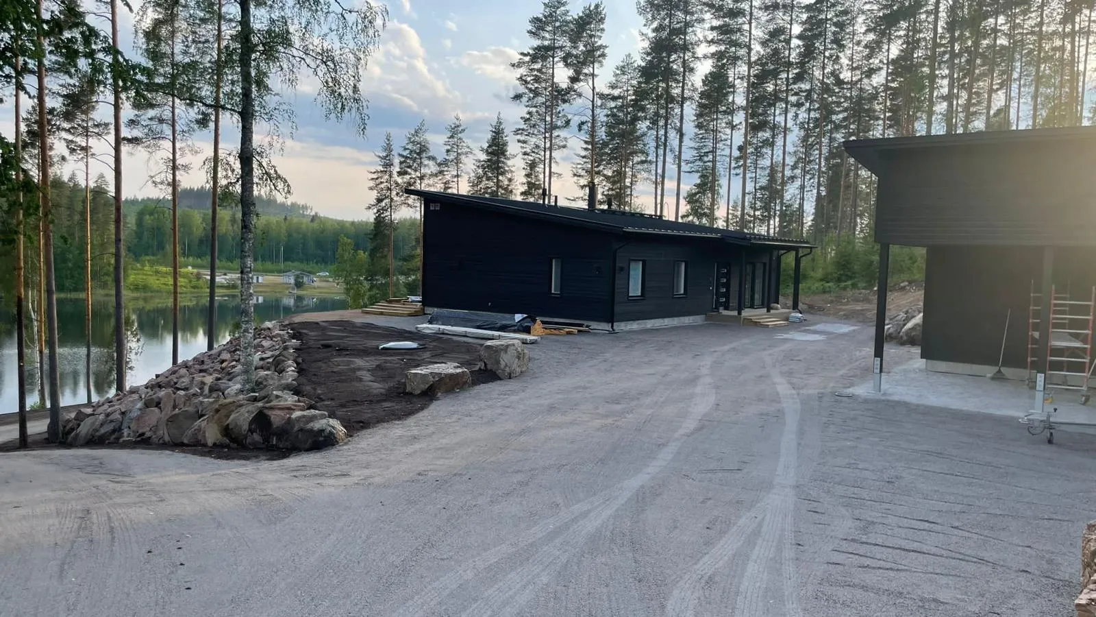 Maisemakuva, jossa näkyy kaksi mustaa taloa järven rannalla, kivikko rannalla ja korkeita puita taustalla, valoisa sää.