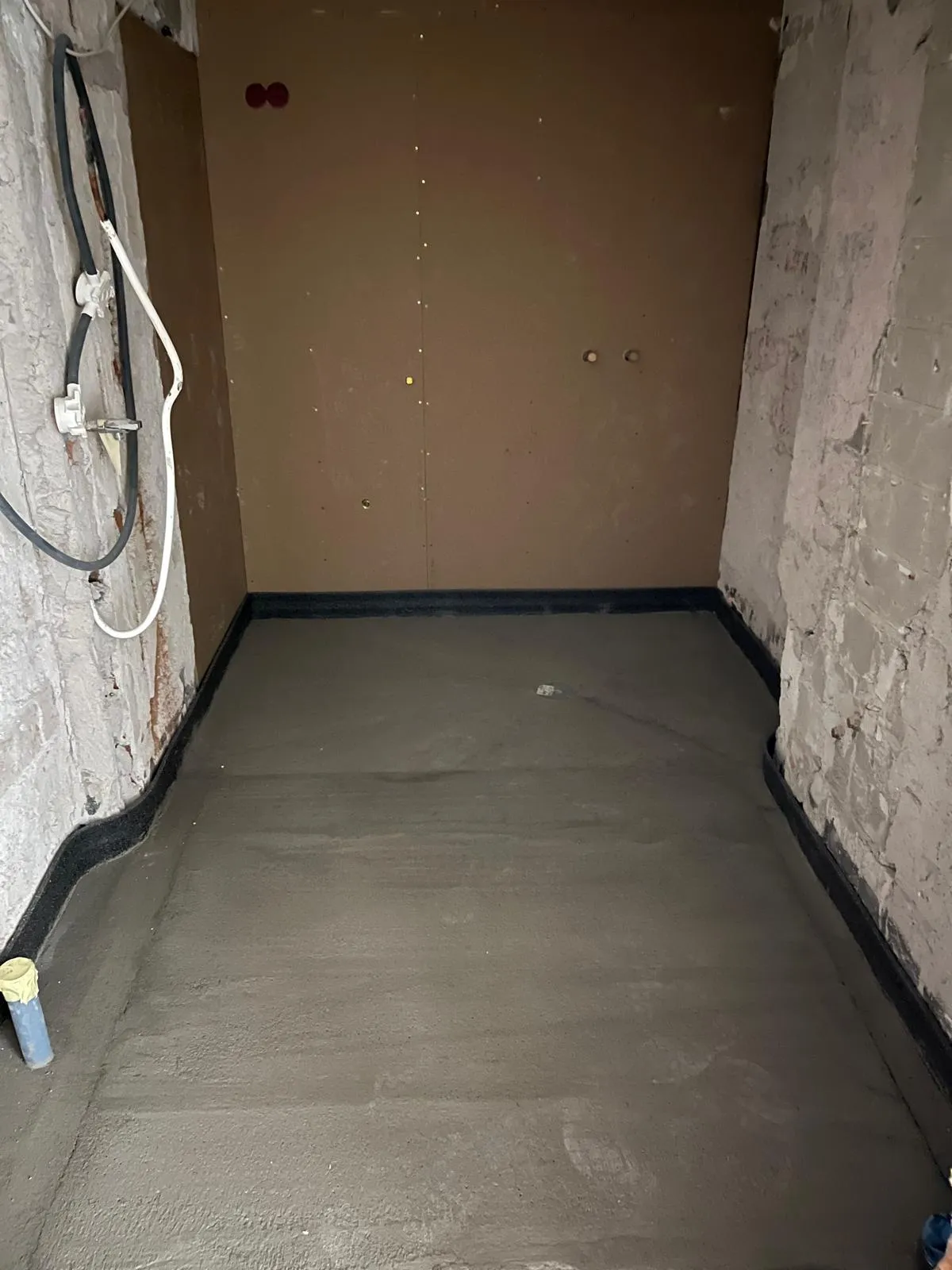 Huoneessa on tasoitettu lattia, jossa on musta reunus, ja seinät ovat osittain rapattuja ja osittain levyjä. Valo on himmeää.