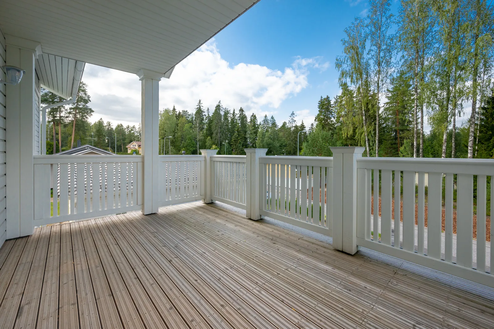 Ulkoterassi valkoisella kaiteella, puulattialla ja näkymällä metsään sinisen taivaan alla.