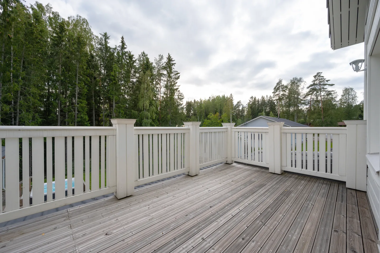Ulkoterassi valkoisella kaiteella, harmaat puulattiat ja vihreä metsä taustalla.