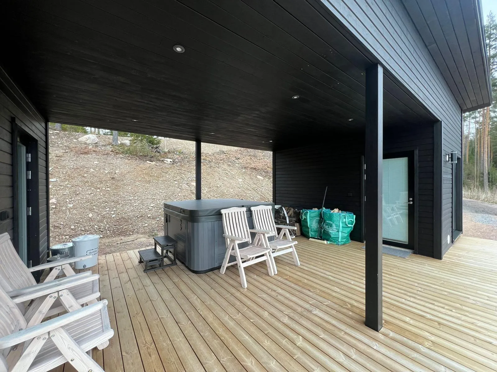 Ulkoterassi, jossa on puinen kansi, harmaa poreallas, kaksi valkoista puutuolia ja kaksi vihreää säkkiä. Terassi on katettu mustalla katolla ja valaistu upotetuilla valoilla.