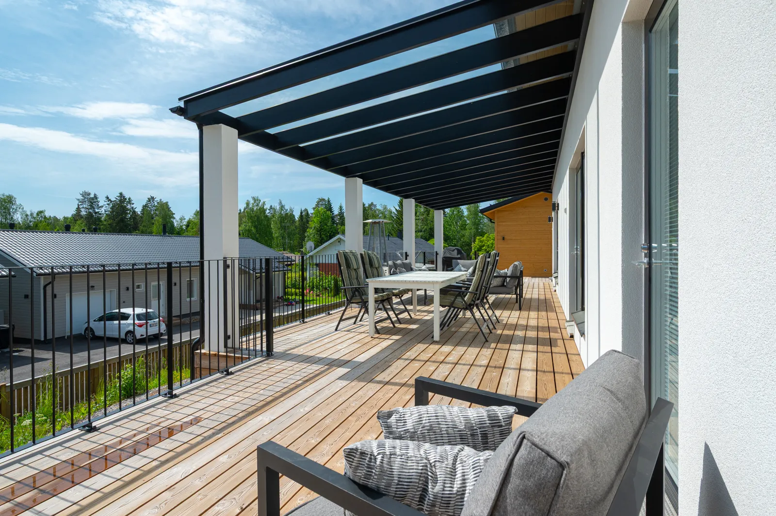 Ulkoterassi, jossa on puukansi, musta metallinen pergola, valkoiset pylväät, ruokailuryhmä ja istuinryhmä. Taustalla näkyy asuinalue. Aurinkoinen sää.