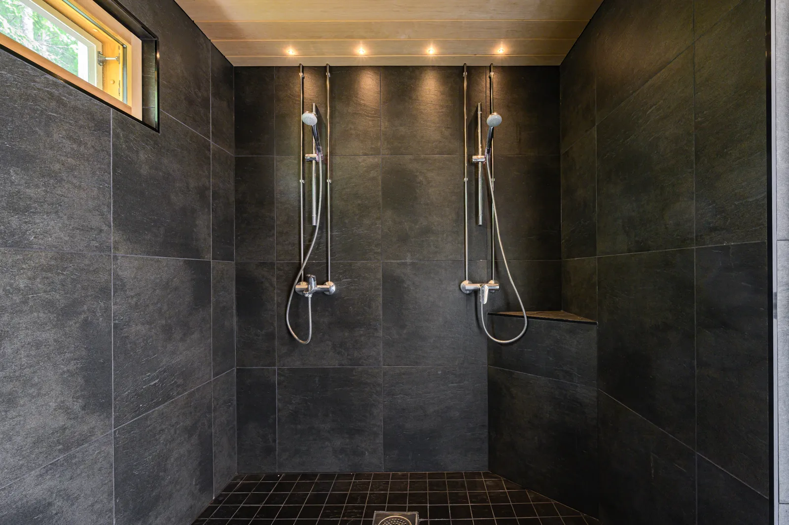 Tumma suihkutila, jossa on kaksi suihkua, mustat laatat seinissä ja lattialla, sekä pieni ikkuna ylhäällä.