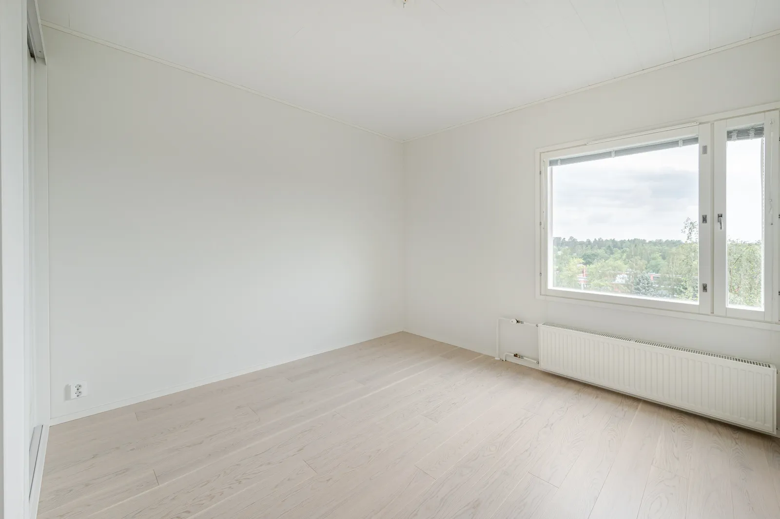 Vaalea, tyhjä huone, jossa on puulattia, valkoinen patteri ja suuri ikkuna, josta näkyy metsäinen maisema.