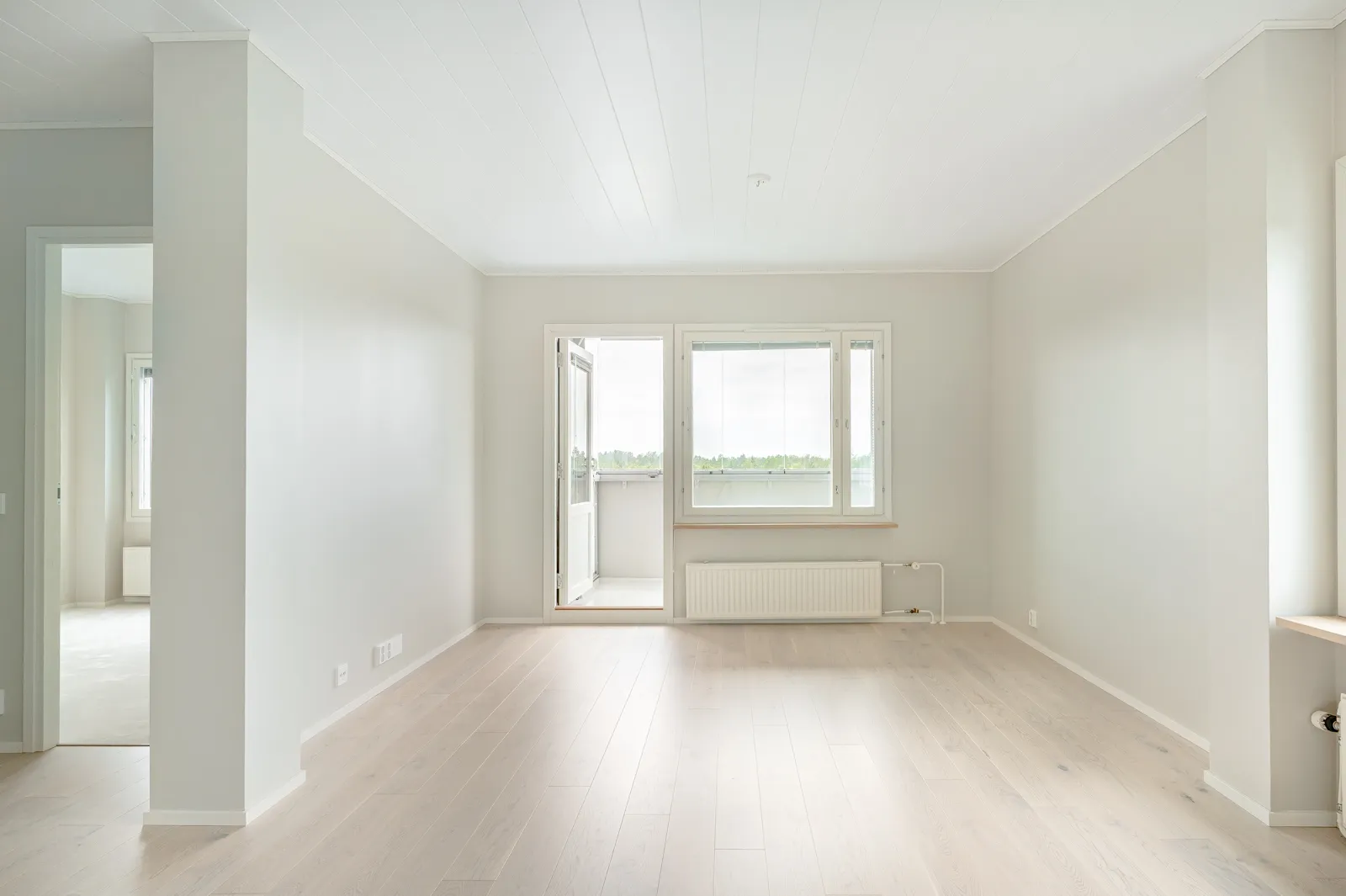 Vaalea, tyhjä huone, jossa on ikkuna ja parvekeovi, sekä vaaleat puulattiat.