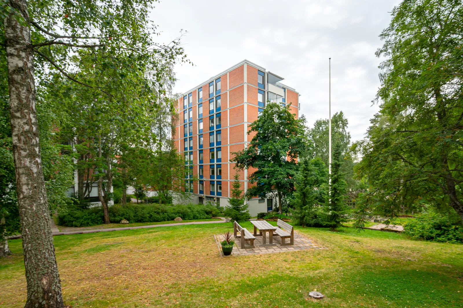 Ulkona on kerrostalo, puita, nurmikko ja piknikpöytä. Rakennus on tiilestä ja siinä on siniset ikkunakehykset.