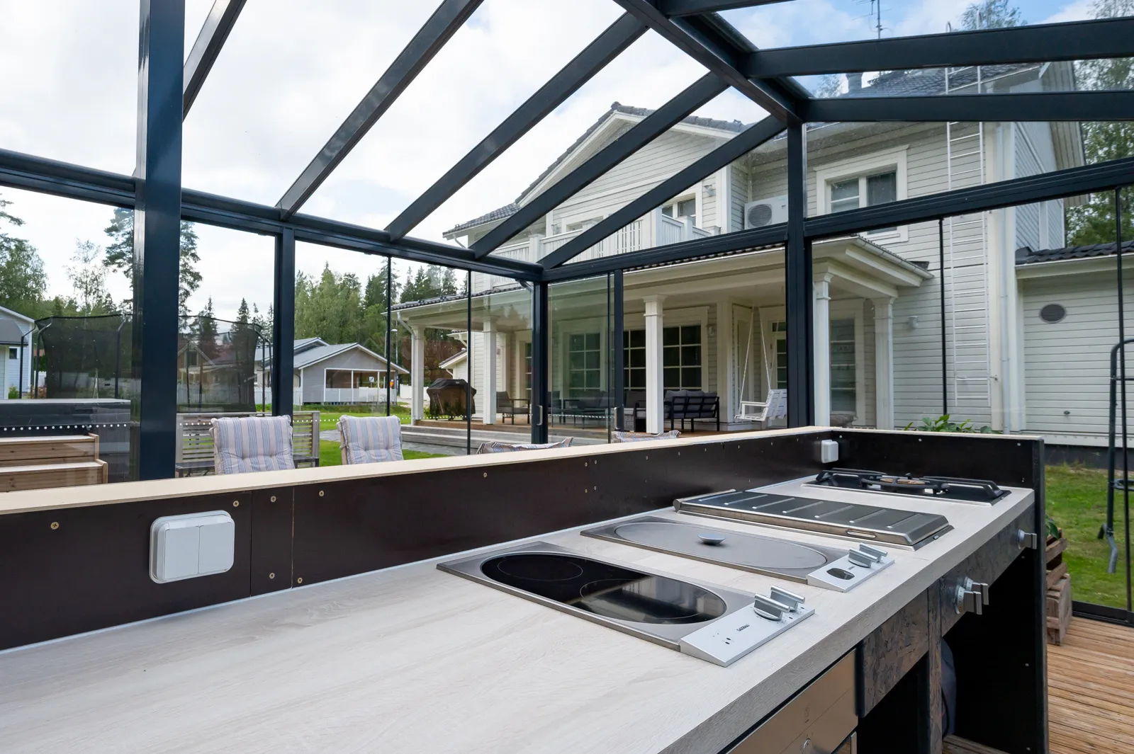 Sisätiloissa oleva ulkokeittiö, jossa on keittotaso, pesuallas ja tumma kehys, taustalla näkyy valkoinen talo ja vihreä maisema.