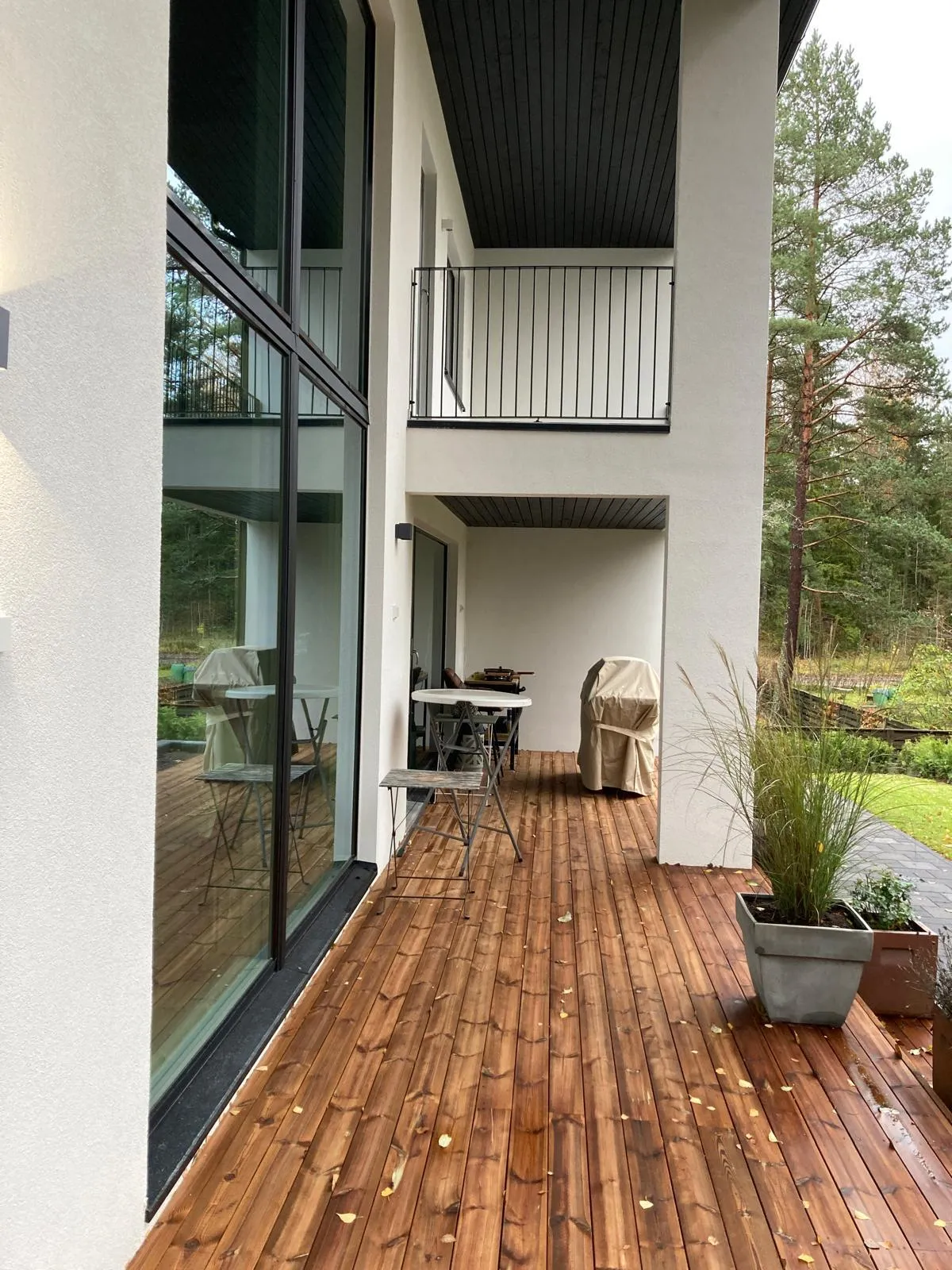 Moderni talon ulkokuva, jossa on puinen terassi, suuret ikkunat ja parveke. Terassilla on pöytä ja tuoleja, sekä kasveja ruukuissa. Valoisa ja avara tunnelma.