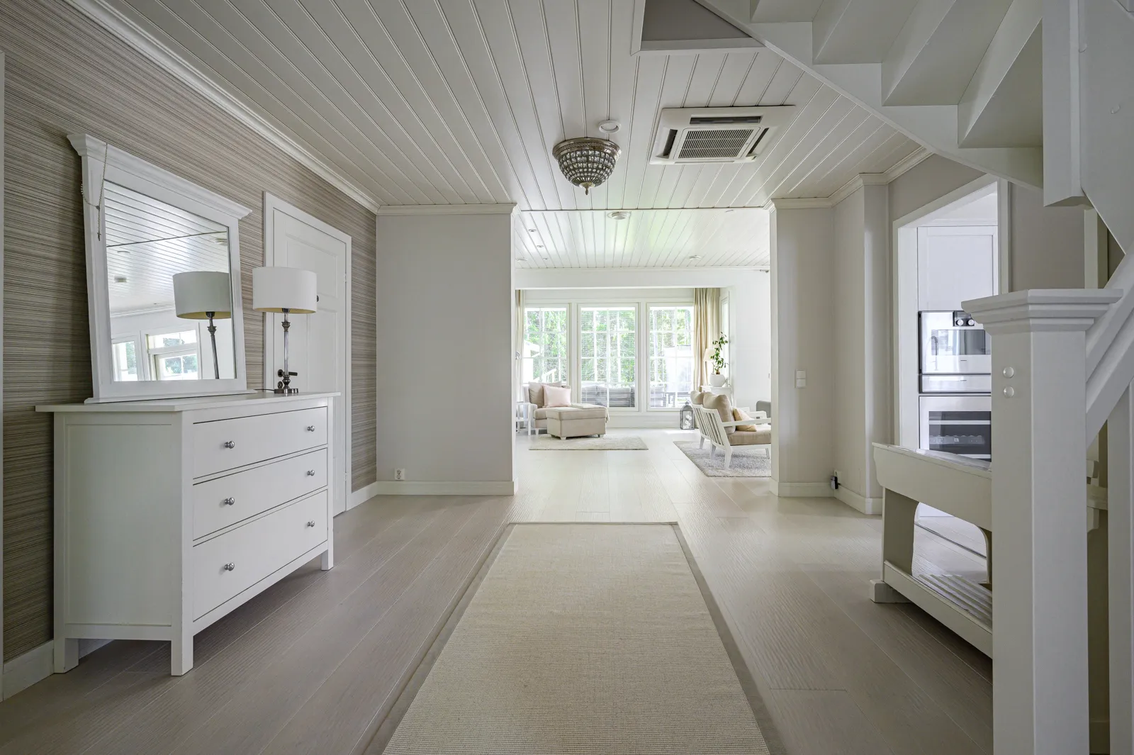 Vaaleassa sisustuksessa on eteinen, jossa on valkoinen lipasto peilillä, matto ja näkymä olohuoneeseen, jossa on ikkunat ja huonekaluja.