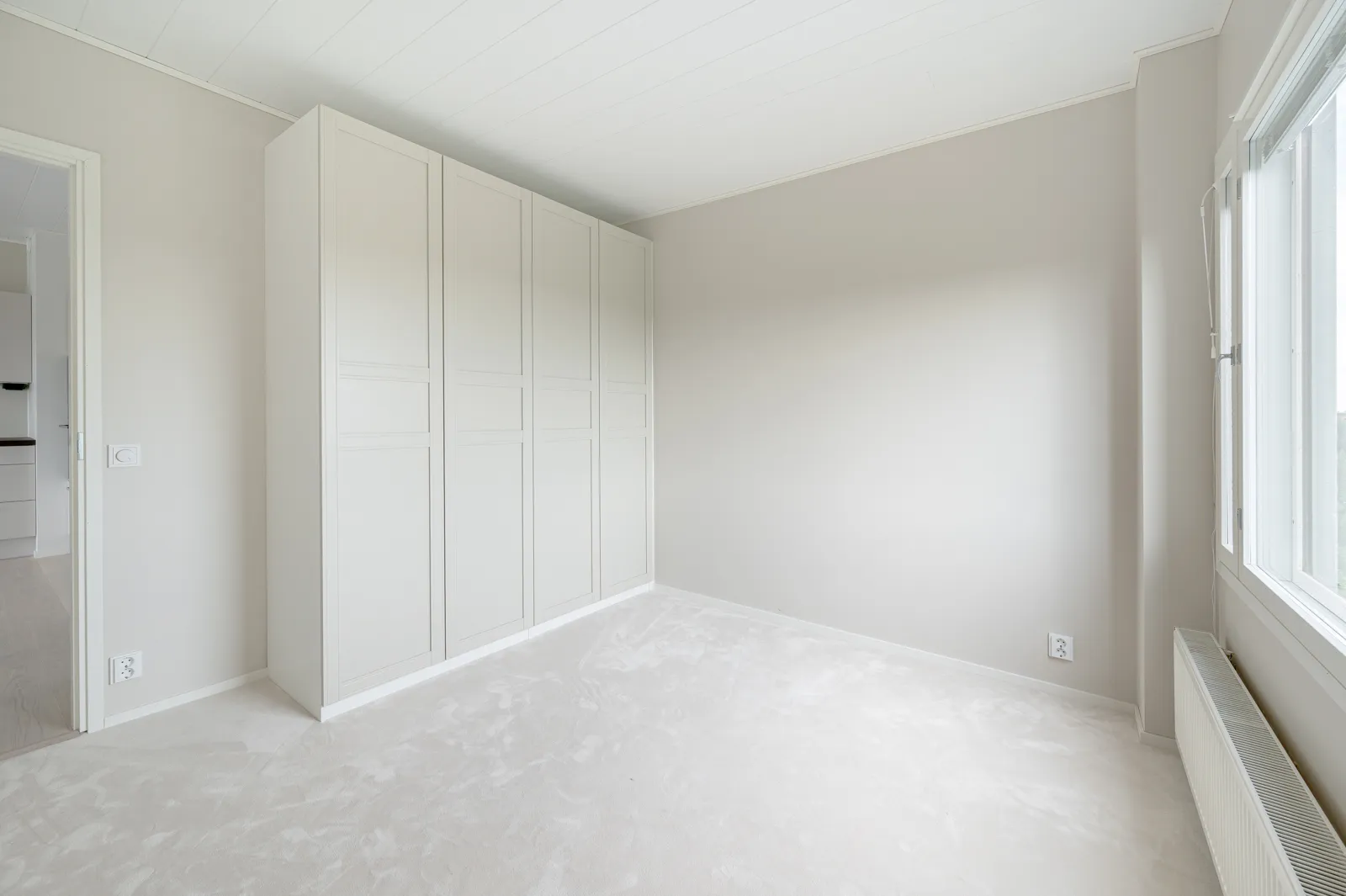 Vaaleassa huoneessa on valkoinen vaatekaappi, ikkuna ja patteri. Huoneessa on valkoinen katto ja seinät.