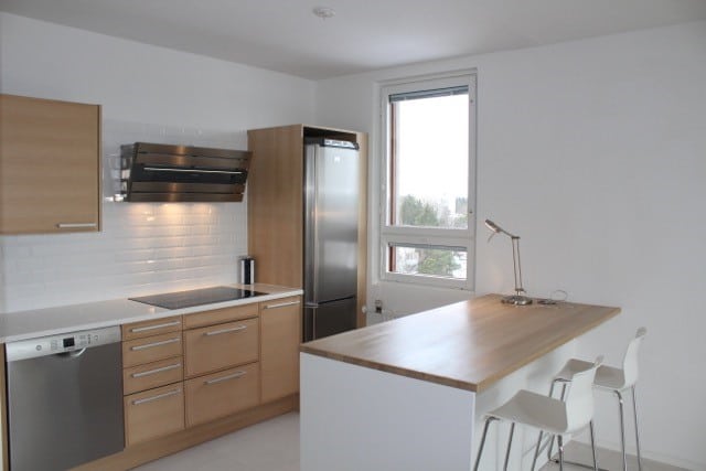 Vaaleassa keittiössä on puukaapit, ruostumattomasta teräksestä valmistettu jääkaappi ja liesituuletin, valkoiset metrolaattat ja keittiösaareke, jossa on kaksi valkoista baarijakkaraa.