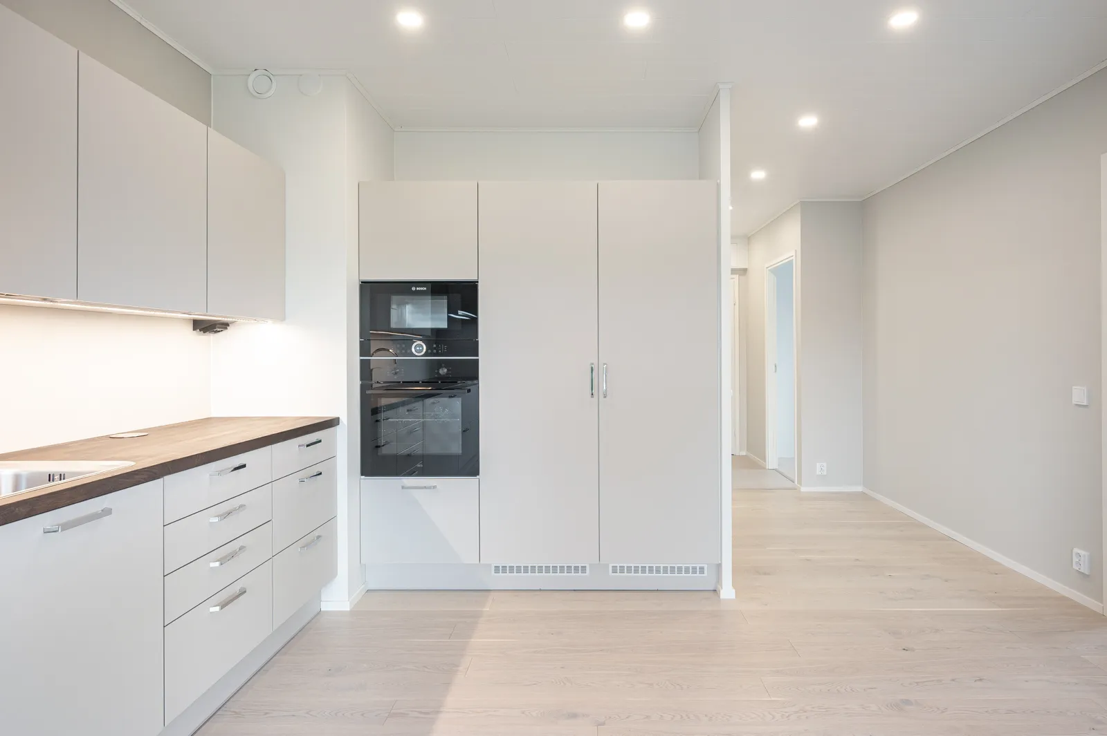 Vaaleassa keittiössä on vaaleat kaapit, tumma työtaso ja integroidut kodinkoneet. Lattialla on vaalea parketti ja katossa on upotettuja valaisimia.