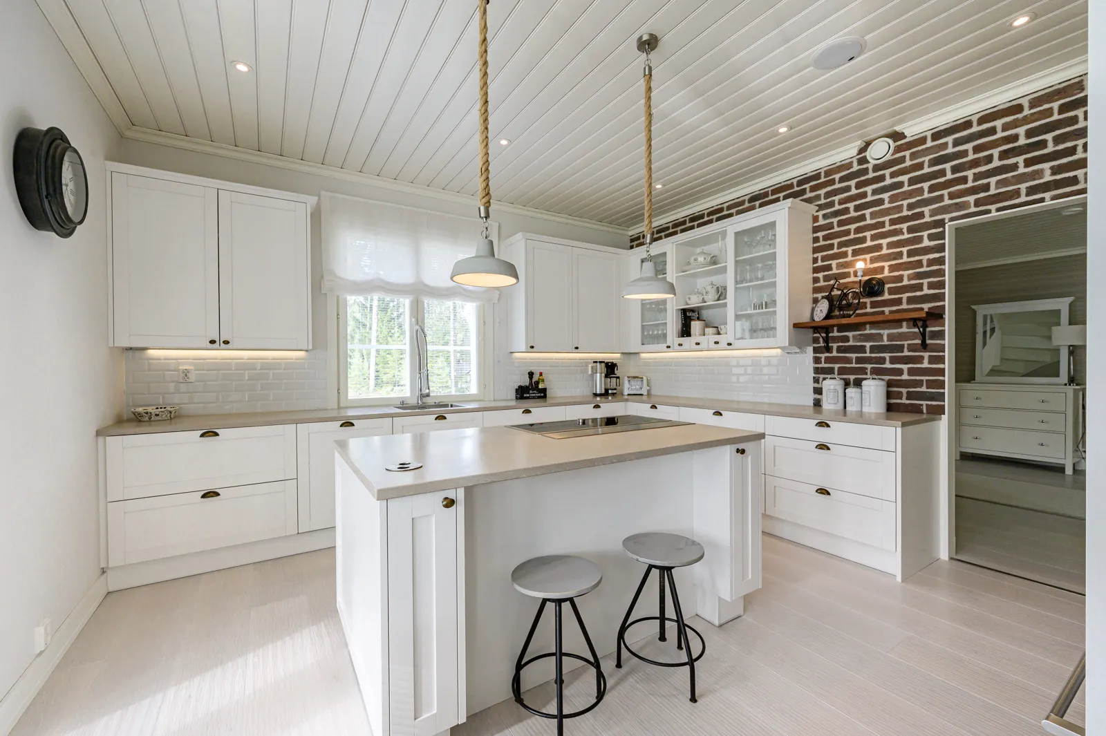 Vaalea keittiö, jossa on valkoiset kaapit, saareke, kaksi jakkaraa, tiiliseinä ja köysivalaisimet.